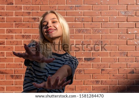 Happy smiling blonde girl in grey striped t-shirt pulls her hands in to the camera isolated over red brick wall, outdoor photo