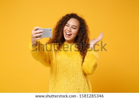 Laughing young african american girl in fur sweater posing isolated on yellow orange wall background. People lifestyle concept. Mock up copy space. Doing selfie shot on mobile phone, spreading hands