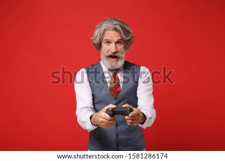 Amazed elderly gray-haired mustache bearded man in classic shirt vest and colorful tie isolated on red background in studio. People lifestyle concept. Mock up copy space. Holding joystick, play game