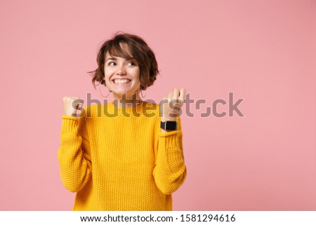 Joyful young brunette woman girl in yellow sweater posing isolated on pink background studio portrait. People lifestyle concept. Mock up copy space. Wearing smart watch on hand, doing winner gesture
