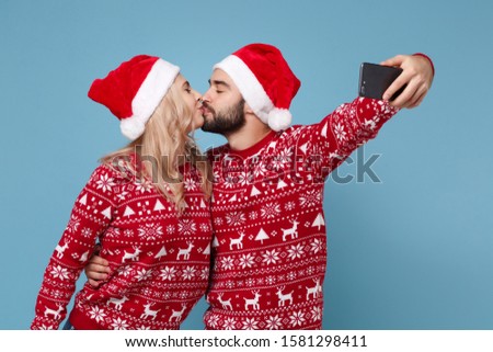 Pretty young couple guy girl in Christmas sweater Santa hat posing isolated on blue background. New Year 2020 celebration party concept. Mock up copy space. Doing selfie shot on mobile phone kissing
