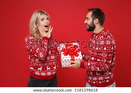 Amazed young couple guy girl in Christmas sweaters posing isolated on red background. Happy New Year 2020 celebration holiday party concept. Mock up copy space. Hold present box with gift ribbon bow