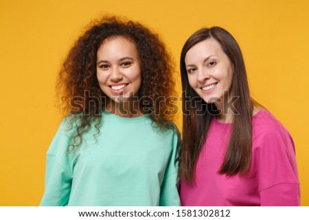 Two smiling beautiful women friends european and african american girls in pink green clothes posing isolated on yellow orange background studio portrait. People lifestyle concept. Mock up copy space