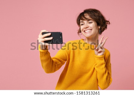 Smiling young brunette woman girl in yellow sweater posing isolated on pastel pink background. People lifestyle concept. Mock up copy space. Doing selfie shot on mobile phone, showing victory sign