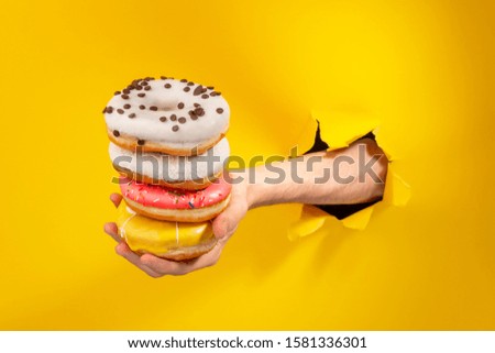 Hand holding a pile of different doughnuts through a torn hole in yellow paper background. Various tastes and glaze. Bakery advertisement.