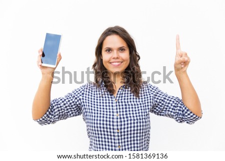 Happy positive mobile phone user showing blank screen and pointing index finger up. Young woman in casual checked shirt standing isolated over white background. Presentation concept