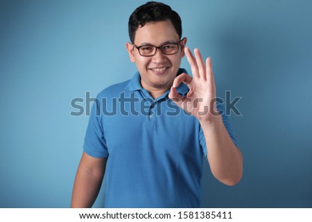 Young Asian man shows okay OK sign. Close up body portrait against blue background