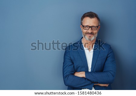 Self-assured attractive middle-aged man standing with folded arms smiling at the camera over a blue studio background with copy space Royalty-Free Stock Photo #1581480436
