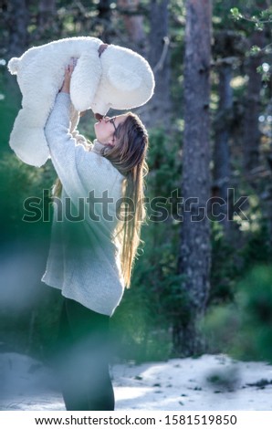 Stock photo of blonde girl lifts the teddy bear high giving it a sweet kiss in the middle of the forest. Sunlight reflects in your hair, concept of happiness, gift.