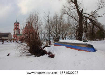 Old overturned wooden boat and damaged tree on snow and Church background, beautiful Russian winter rural landscape