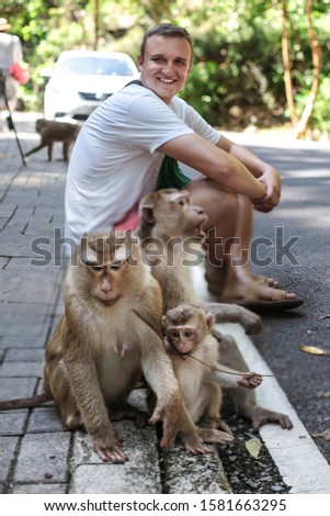 Young attractive man seats with a monkey family and smiles. Cute monkey family with a little baby in Thailand, Phuket, Monkey hill.