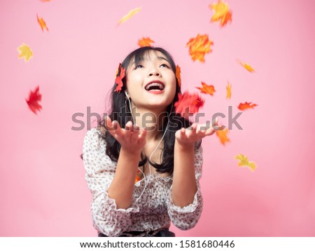 Happy Asian teenager girl wearing using earphone and playing with dropping maple leafs on pink background.