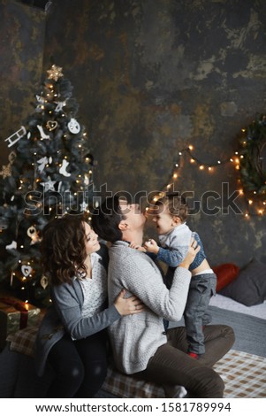 Young parents and the baby boy hugs together in the interior decorated for New year. The father, mother and their little son have fun in the Christmas interior. Happy New Year and Merry Christmas