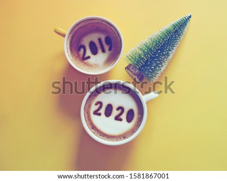 Goodbye 2019, Hello 2020 theme coffee cup with number 2020 on frothy surface and another one with 2019 at the bottom of cup over yellow background with Christmas tree. Holidays food art concept.