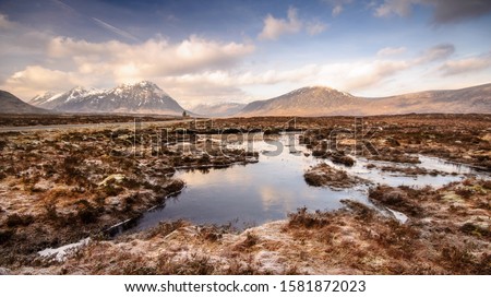 Lochan ponds are frozen over with winter ice on the vast wetland peat bog landscape of Rannoch Moor, with Buachaille Etive Mor and the mountains of Glen Coe rising in the distance. Royalty-Free Stock Photo #1581872023