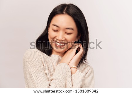 Calm brunette female with closed eyes, keeps both palms on heart, feels gratitude. young woman Asian appearance dressed in knitted warm sweater stands isolated white background in Studio. 