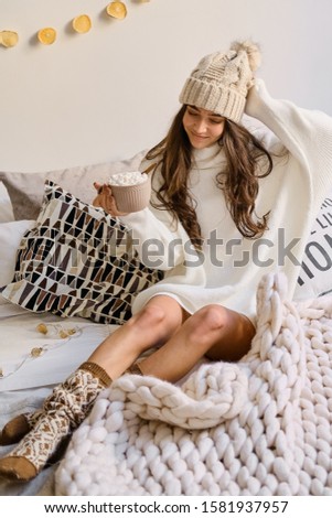 A cheerful  caucasian girl in a hat and sweater sits on the bed with a cup of hot chocolate with marshmallows. Christmas is all in the home.
