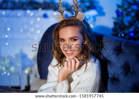 A large portrait of a girl with a broad smile and deer horns on her head sits on a sofa at home. Christmas Eve. New Year.