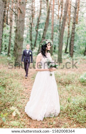 Gorgeous bride in floral wreath holding bouquet, standing in forest. Handsome groom is going to his bride on the background. Rustic wedding outdoors