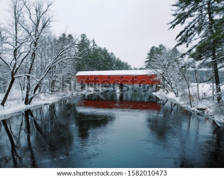 Beautiful aerial drone view looking up river towards old-fashioned red covered bridge after a fresh snow fall Royalty-Free Stock Photo #1582010473