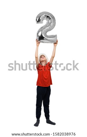 Boy in a Santa hat is holding a silver inflatable number 2 above his head. White background. New Year concept