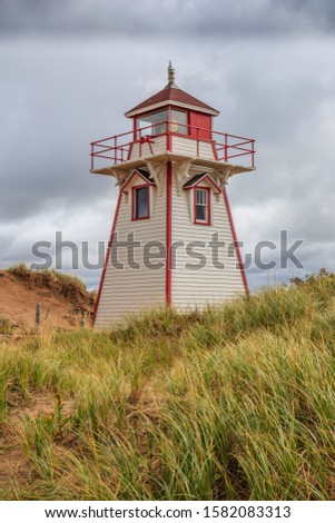 A classic, 19th century, white and red wooden lighthouse shines its light high above grassy sand dunes under dark storm clouds on the Atlantic coast.