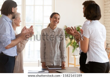 Happy diverse teammates gathering near table, clapping hands, congratulating smiling mixed race young female employee with birthday, business achievement, successful contract signing with client.