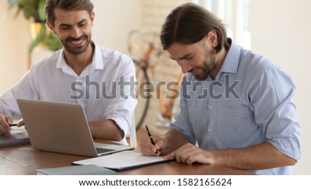 Successful young businessman watching smiling partner signing contract, making commercial agreement after project negotiations. Satisfied with good deal happy client putting signature on document.