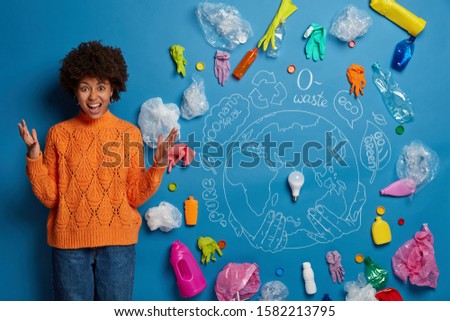 Irritated dark skinned curly woman recycles plastic wastes, yells with anger, keeps hands raised, being frustrated and crazy, overwhelmed by ecology problem, annoyed by pollution of environment