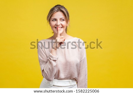 Portrait of cheerful beautiful girl with blonde hair in casual beige blouse standing, looking away with toothy smile and showing silence gesture, indoor studio shot, isolated on yellow background