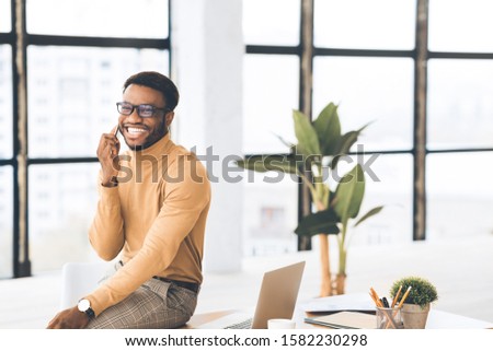 Work Call. Happy african american manager talking on cell phone, sitting on desk. Free space