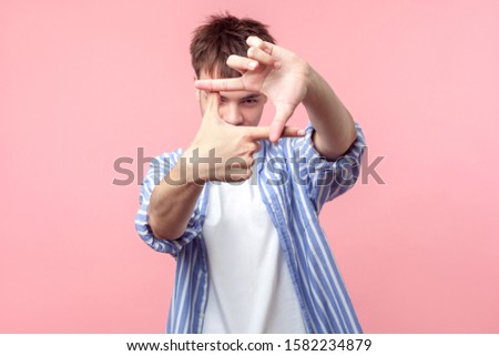 Frame of fingers. Portrait of young man with brunette hair in casual striped shirt looking at camera through hand frame taking picture, focusing the eye. indoor studio shot isolated on pink background