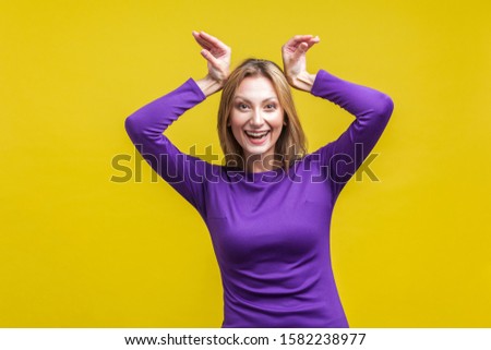 Portrait of adorable funny woman in elegant purple dress showing bunny ears with hands on her head and looking at camera with childish toothy smile. indoor studio shot isolated on yellow background
