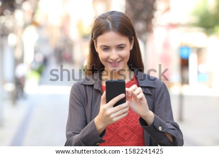 Front view portrait of a happy teen checking mobile phone content walking in the street