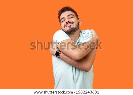 I love myself! Portrait of egoistic brunette man with beard in white t-shirt standing with closed eyes, embracing himself and smiling form pleasure and proud. indoor, isolated on orange background Royalty-Free Stock Photo #1582243681