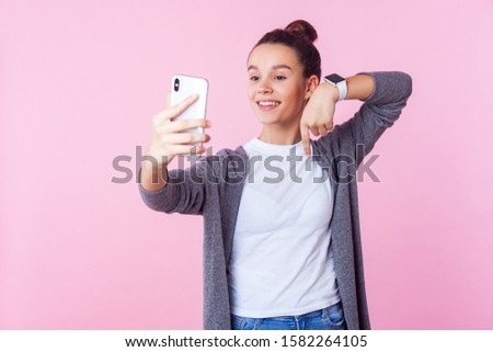 Subscribe here! Portrait of positive blogger, teenage girl with bun hairstyle in casual clothes smiling and pointing down while streaming online using phone. studio shot isolated on pink background