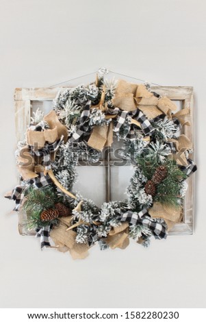 Flocked homemade country or hunters Christmas wreath over rustic farmhouse window on a grey wall. Made pine cones, fir branches, buffalo plaid ribbon, burlap and deer antlers. Flocked with fake snow.