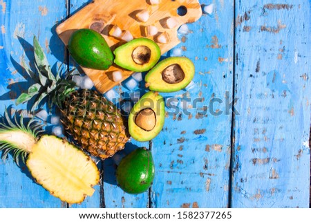 Summer composition with tropical fruits avocado and pineapple, cutting board and ice cube on blue wooden color background