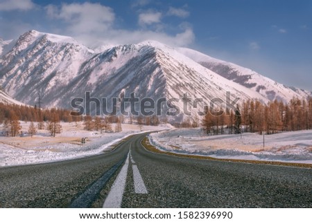 Picturesque winter landscape with the asphalt road in the mountains covered with snow and trees on the side of the road on the background of the blue sky and clouds
