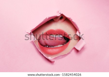 Girl bites her tongue, tongue piercing. Big beautiful lips, piercing parlor. Beautiful teeth, dentistry. Pink background, girl's face through torn cardboard. Royalty-Free Stock Photo #1582454026