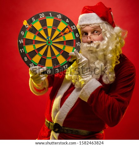 Santa Claus holds a darts target in his hands and poses on a dark red background