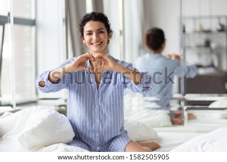 Happy smiling woman looking at camera and showing love sign with hand while sitting in bed with white bedding home portrait. Beautiful cheerful loving lady in nightgown making heart shape with fingers