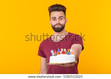 Embarrassed young male hipster with beard holding a birthday cake in his hands and looking thoughtfully at him posing on a yellow background. Concept of time quickly flies by.