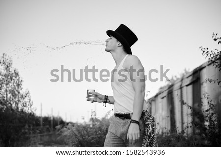 Three-quarter portrait of young man, wearing grey pants,white top, black classic hat, holding water glass, spitting on the side. Black and white picture of creative man on abandoned construction site.