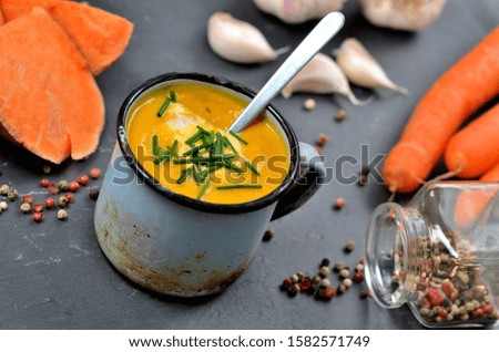 Sweet potato soup with cream and fresh chive in old enamel cup. Garlic, pumpkin and half pieced carrot in background. Slate plate and spoon.