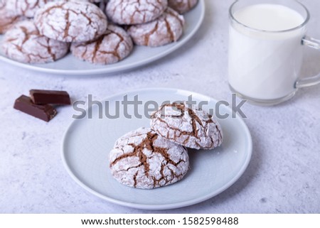 Chocolate cookies with cracks, strewed with icing sugar. Homemade baking. Background: a cup of milk, chocolate and a plate of cookies. Selective focus, close-up.