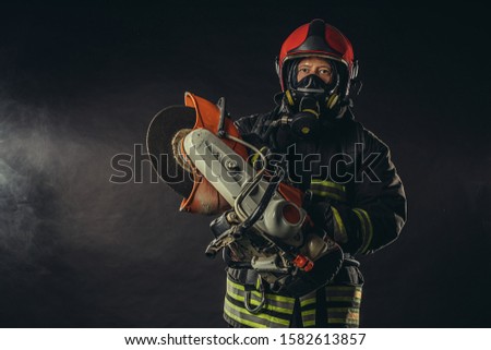 young extinguisher holding chainsaw stand isolated over smoky background, wearing fireman suit, confident firefighter