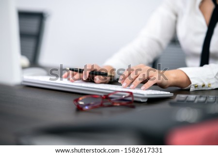 Women in white shirt and black ties is typing on white keyboard. Calculator files and mono-block computer is on the desk. Women holding black pencil with right hand.