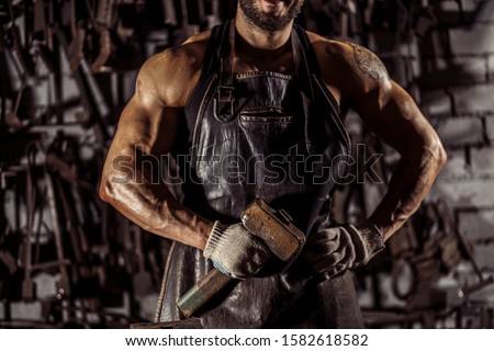 cropped man with muscular body, young forger hold hammer in hands, have beard. instruments in the background