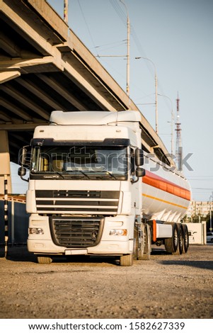 Truck with trailer, tank with flammable liquid, under a large bridge at the pier on the river bank, sunset light, white and red cars standing on gravel